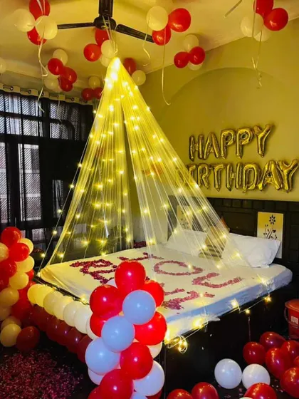 A romantic birthday decoration for a partner. The setup includes a fairy light canopy, "Happy Birthday" foil balloons, and red and white balloons creating a festive and intimate atmosphere.