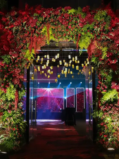 A floral archway of red and green botanicals creates a vibrant transition into the main event space, offering a glimpse of the dazzling party inside.