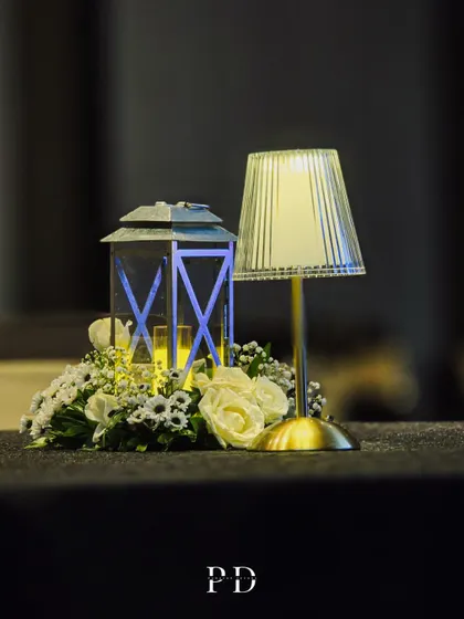 A simple yet elegant centerpiece for a cocktail event, featuring a modern lamp, a classic lantern, and a small arrangement of white roses.