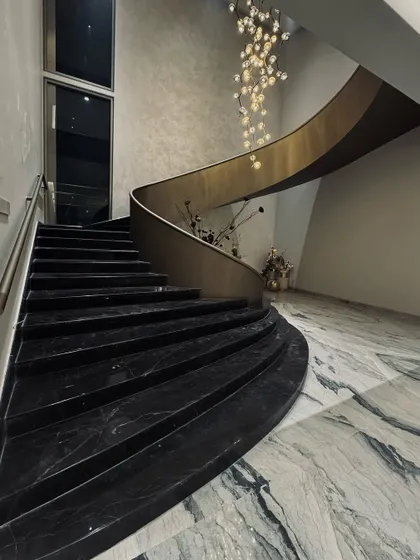 This view of the sculptural staircase shows its elegant curve and the contrast between the dark marble steps and the light marble flooring. A cascading chandelier follows the sweep of the stairs, adding a touch of glamour and illuminating the entire entryway.