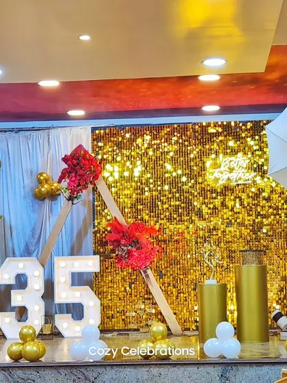 A stage setup for a 35th birthday, featuring a gold sequin wall, marquee numbers, and professional lighting umbrellas for a photo-shoot feel.