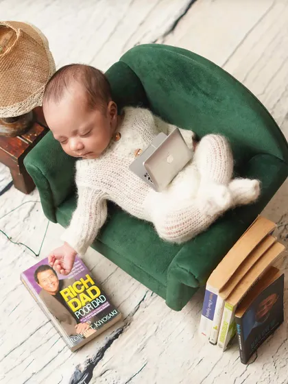 CEO of midday naps! This humorous setup features a newborn sleeping on a miniature couch with a tiny laptop and a copy of 'Rich Dad Poor Dad'.