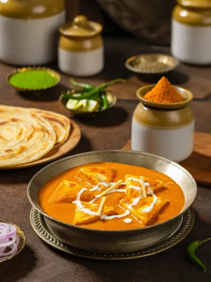 Shahi Paneer in a traditional metal bowl, surrounded by pickling jars and parathas. This composition for Rakesh Masala creates a feeling of a complete, wholesome meal.