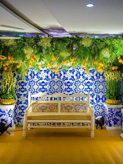 A unique Mehendi seating area with a backdrop of blue and white Portuguese-style tiles. The vibrant yellow and blue floral arrangements create a stunning contrast, perfect for a themed celebration.