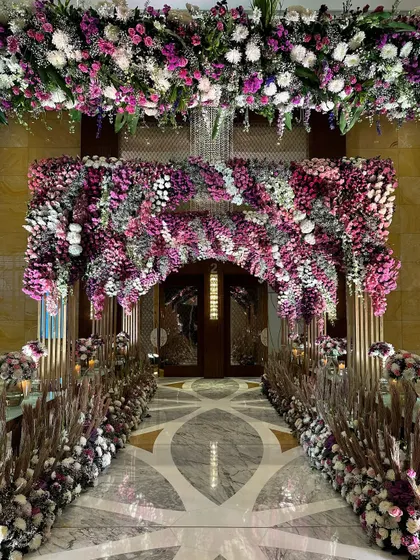 A straight-on view of the magnificent floral entrance. The archway, thick with purple and white flowers, perfectly frames the doorway, inviting guests into a world of celebration.