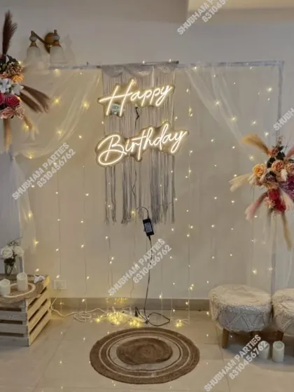 A stylish and modern boho-themed birthday decoration perfect for an intimate celebration. This setup features a macrame wall hanging, a "Happy Birthday" neon sign, fairy lights, and dried floral arrangements for a chic and rustic vibe.