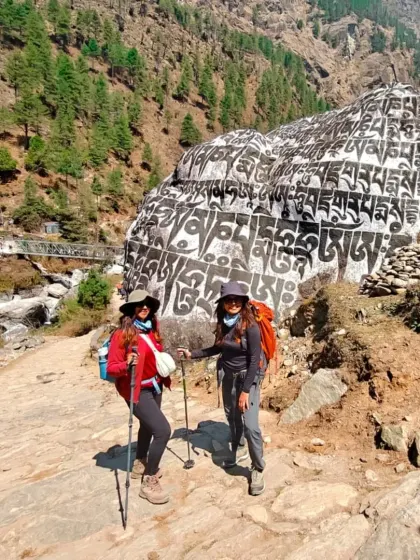 Two trekkers stand before a large mani stone inscribed with Buddhist prayers. The trail to EBC is not just a physical journey but a spiritual one, steeped in the local Sherpa culture.