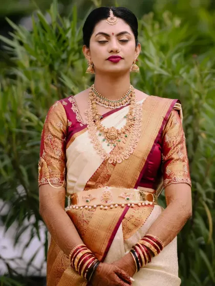 A serene portrait of a bride in a classic off-white and maroon silk saree. Her makeup is bold and traditional, with a deep berry lip.