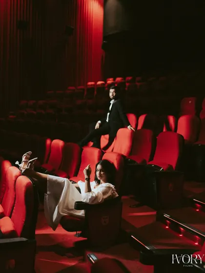 A stylish, full-length shot of the couple owning the theatre space. Her relaxed pose with feet up and his confident stance on the seat back create a playful, high-fashion image that tells a story of a modern power couple.