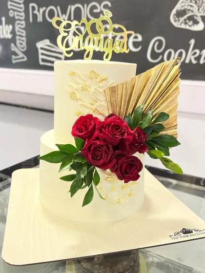 A chic two-tiered engagement cake with a crisp white finish. The design is elevated with a bold arrangement of fresh red roses, a textured white chocolate sail, and a modern gold palm leaf.