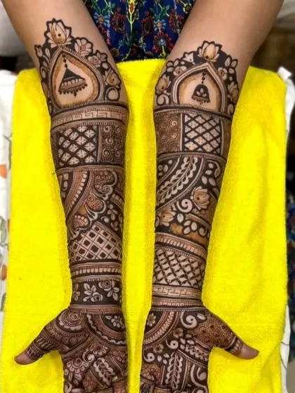 A bridal design featuring hanging bells (jhumkas) at the top, with a mix of geometric and floral patterns covering the full arm.