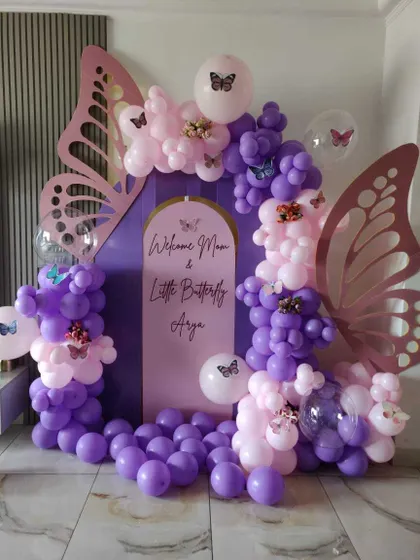 A 'Welcome Mom & Little Butterfly' baby shower theme. An arched purple backdrop is framed by a purple and pink balloon garland with large butterfly wing cutouts.