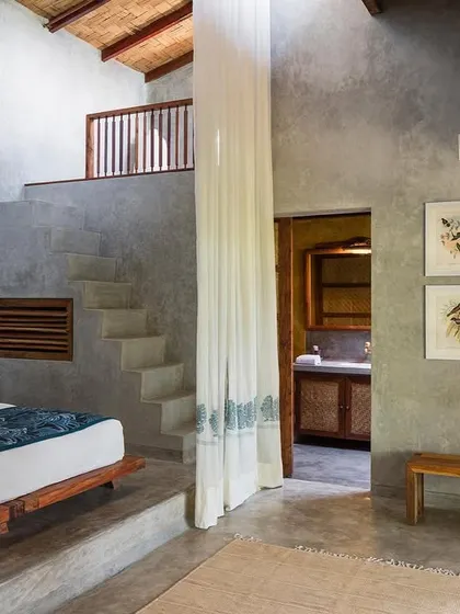The main bedroom area of the Blue Room, with stairs leading up to the mezzanine. The space is characterized by its use of raw concrete and natural textures.