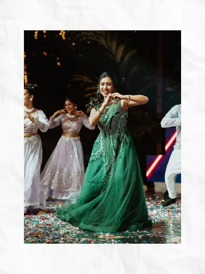 The bride makes a heart shape with her hands as she performs a dance at her Sangeet, surrounded by backup dancers.