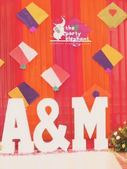 A fun and colorful photo backdrop for the Mehendi, featuring a wall of kites and the couple's initials, set against vibrant orange and pink drapes.