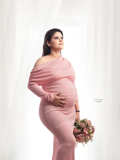 A classic three-quarter maternity portrait. The client looks radiant in a pink off-shoulder gown, holding a matching bouquet against a bright and airy studio background.