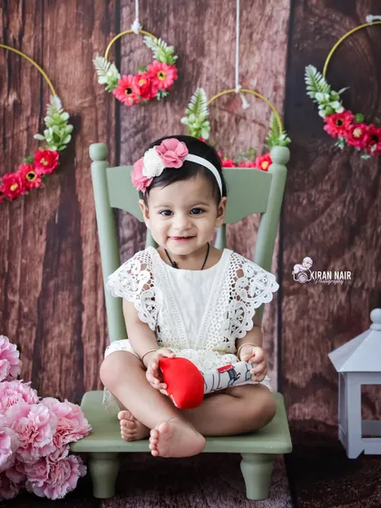 A sweet toddler girl in a white lace dress sits on a small chair in a rustic-themed studio setup. The wood-paneled background and floral hoops add a charming touch.