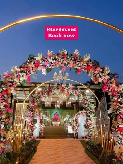 A luxurious wedding entrance at dusk. The walkway is lined with multiple floral arches in pink and white, illuminated by warm lights and golden pillars, creating a magical tunnel effect.