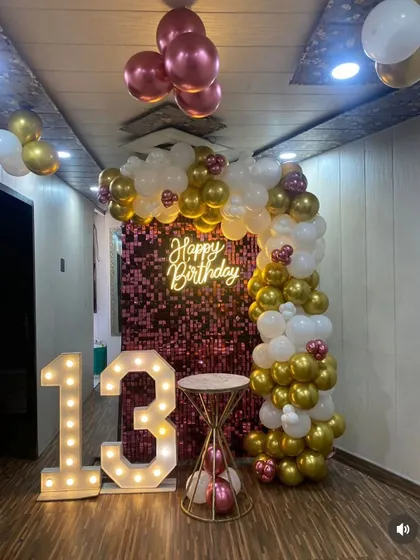 A stylish 13th birthday decoration with a pink shimmer wall. The setup includes a white and gold balloon arch, a neon sign, and large marquee numbers.