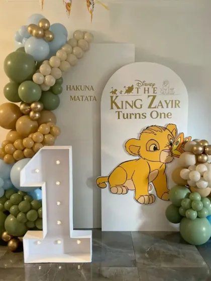 Hakuna Matata! A sweet Lion King theme for a first birthday celebration. This setup includes a baby Simba cutout, a marquee number "1", and a balloon garland in gentle greens, blues, and browns.