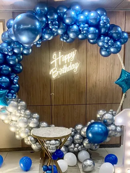 A stylish birthday photo booth with a circular balloon frame in chrome blue and silver. The setup is completed with a "Happy Birthday" neon sign and a light-up number '1'.
