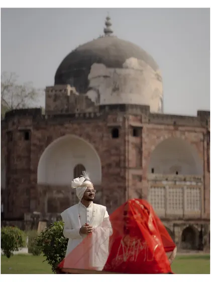 An artistic shot with the groom in focus and the bride, veiled in red, creating a beautiful, soft silhouette against the backdrop of a historic tomb.