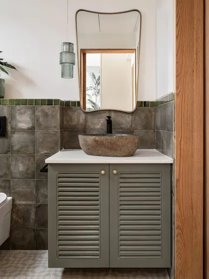 The powder room has an "airy, beachy, moody" vibe. We used handmade-look grey-green tiles, a natural stone sink, and a louvered vanity to create a space with tons of texture and character.