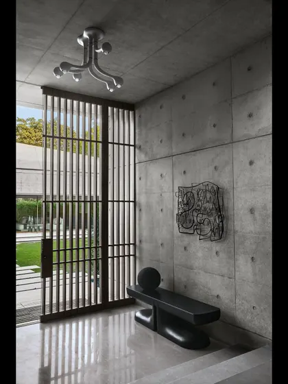 A quiet corner in the entrance vestibule, where sculptural furniture and art are placed against the textured concrete wall. The vertical wooden screen provides a filtered view of the garden beyond.