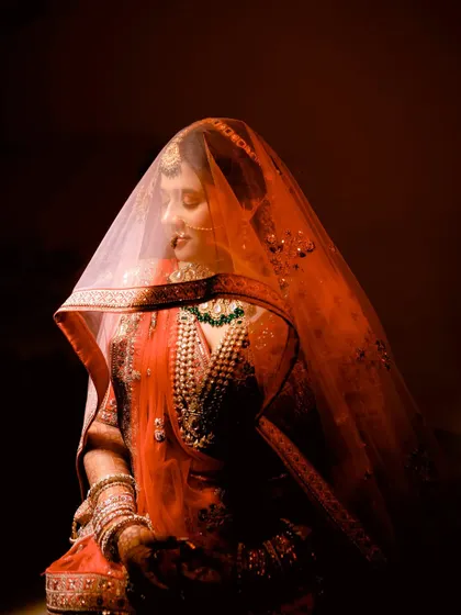 A stunning bridal portrait with dramatic lighting. The bride is veiled and looks down, creating a sense of serenity and grace. The rich red tones make this a powerful and artistic image.