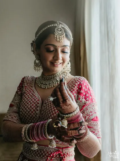 A classic bridal portrait of Shivani, smiling as she puts on her bangles, bathed in soft window light.