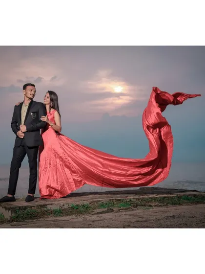 A dynamic and romantic pre-wedding shot on the coast, with the wind catching the bride's long gown, creating a sense of movement and drama against the sunset.