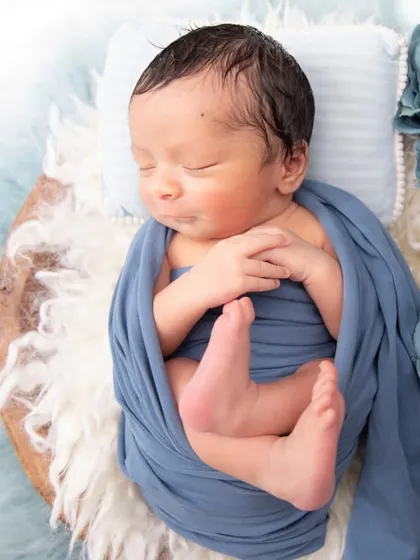 A newborn wrapped in a dusty blue swaddle, with his tiny feet peeking out. The addition of blue roses adds a soft, artistic touch.