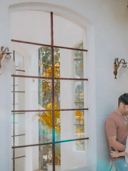 An intimate portrait of a couple against a large, elegant window. The natural light and surrounding foliage create a soft and romantic mood for this pre-wedding photo.