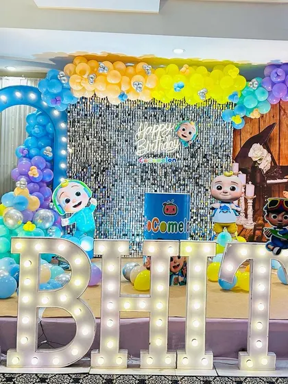A Cocomelon-themed party with a silver shimmer wall and a "Happy Birthday" neon sign, surrounded by colorful characters and balloons.