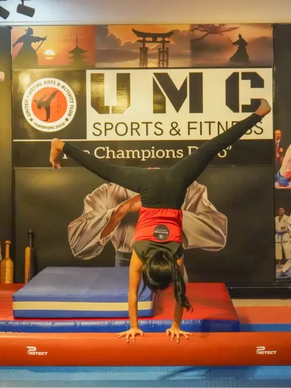 A gymnast holds a perfect handstand split on the balance beam, a move that requires immense strength and balance. Our UMC logo in the background represents the supportive environment where such skills are honed.