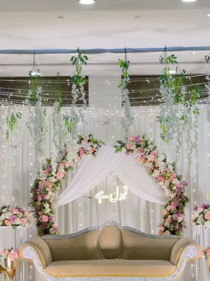 A dreamy Nikah backdrop with a floral arch and draped canopy. The decor features hanging wisteria and a 'Qubool Hai' neon sign, creating a soft and romantic ambiance.