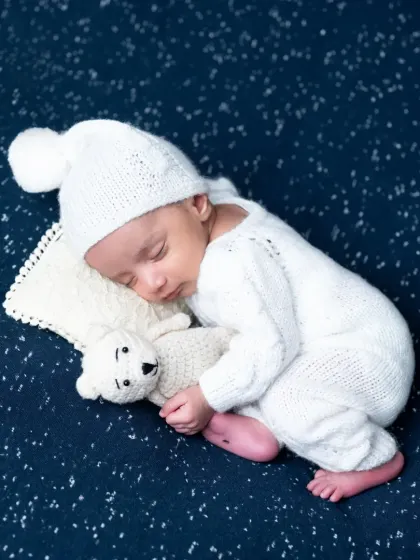 A newborn boy in a white knit outfit and pom-pom hat, cuddling a small teddy bear as he sleeps on a starry blue background.
