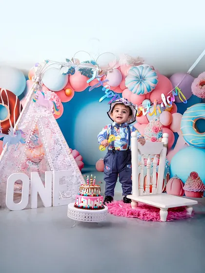 A one-der kid in the house! This little boy is all smiles for his first birthday, standing proudly in a colorful setup complete with a teepee, balloons, and a delicious-looking cake.