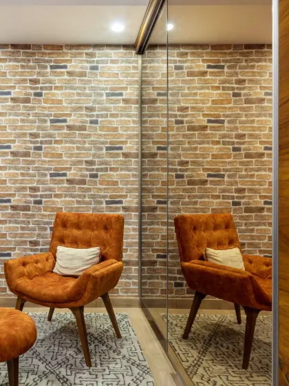 The client's favorite lounge chair in a vibrant orange finds its home in the den. Paired with an ottoman, it creates a perfect haven for literary retreats against the rustic brick wall.