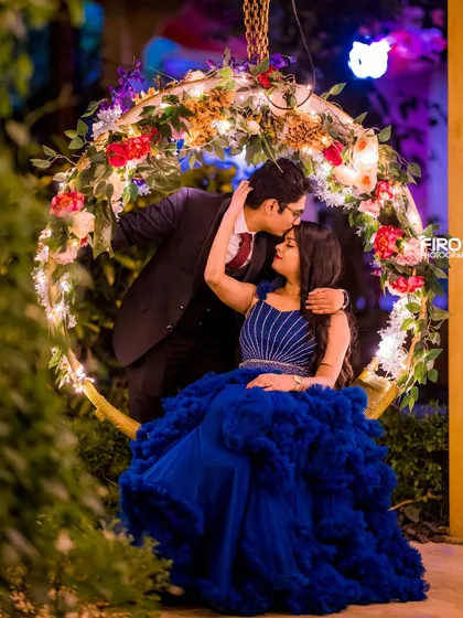 A tender moment on the floral swing. The groom-to-be kisses his partner's forehead, while her ruffled blue gown adds texture and romance to this magical night shot.