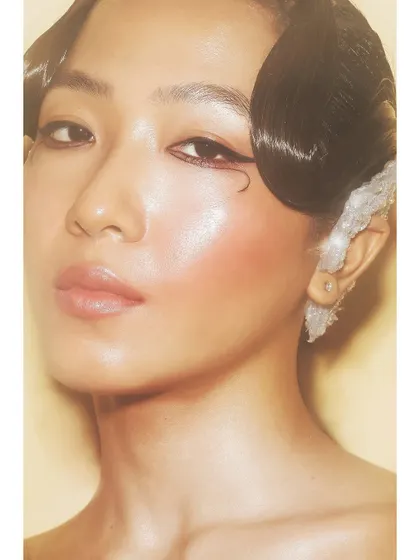 A close-up beauty shot with a warm, golden glow. The focus is on the model's flawless skin and the delicate details of her makeup and hair.