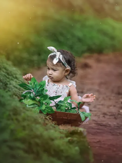 A toddler sits in a wooden bowl, looking thoughtfully at the leaves. The warm sunlight and magical dust motes create a dreamy, fairytale atmosphere.