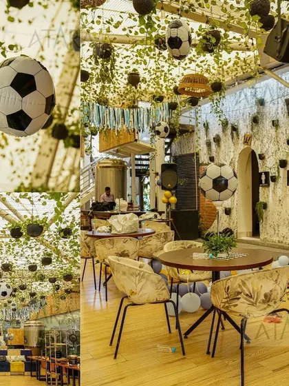 A wide view of a soccer-themed party held indoors. The ceiling is covered with green vines and hanging soccer balls, transforming the space into a lush, green soccer field.