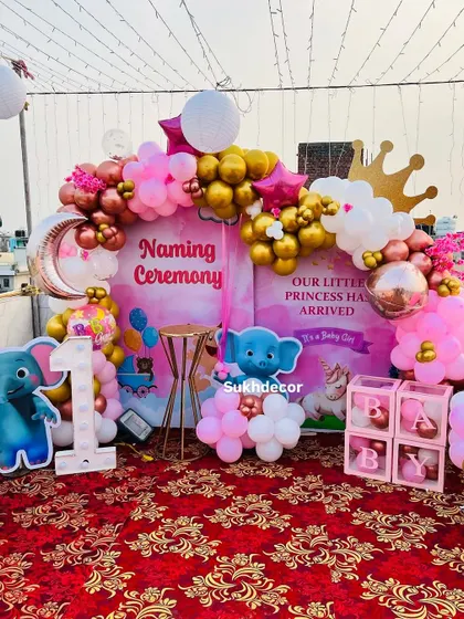 A clear, centered shot of the naming ceremony backdrop. The design includes a crescent moon balloon, a crown accent, and "BABY" blocks, all tied together with a pink and gold color scheme.