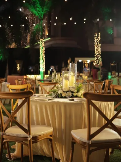 An outdoor dining area with wooden cross-back chairs and simple, elegant table settings. The palm trees wrapped in fairy lights add a magical touch to the natural surroundings.