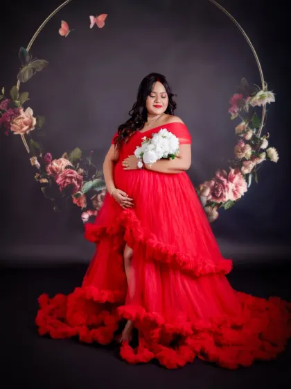 A stunning solo portrait with a floral hoop backdrop. The mom-to-be holds a white bouquet, looking serene and beautiful in her vibrant red gown.