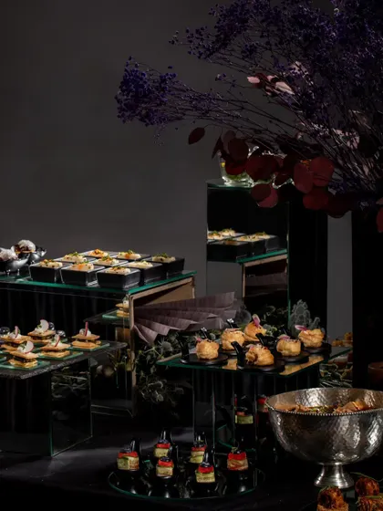 A wide shot of a sophisticated appetizer station at a Diwali party. The use of mirrored stands and dark florals creates a chic and modern festive ambiance.