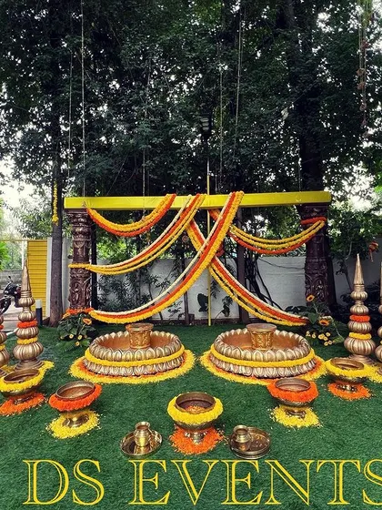 An elaborate outdoor Haldi or Mehendi setup. The decor includes draped floral garlands, traditional brass urlis for the ceremony, and decorative spires, all set on a lush green lawn.