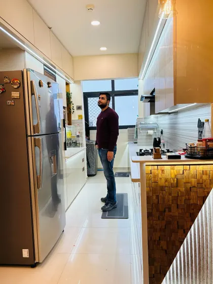 Standing in a newly finished parallel kitchen. The design features a mix of white and wood-textured laminates, a large refrigerator, and a breakfast bar with a wooden block design, making it both modern and warm.