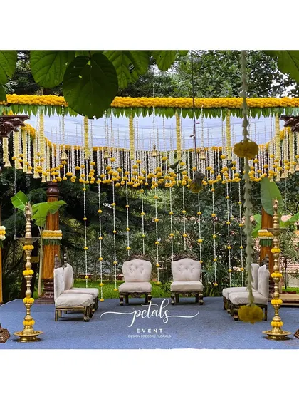 A closer look at the South Indian wedding mandapa, framed by lush greenery. The design features wooden pillars, yellow marigolds, and leaf parrots, celebrating our rich cultural heritage.